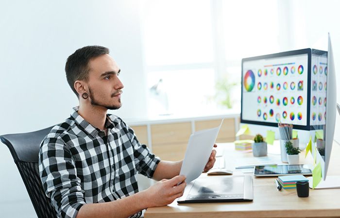 Grafiker sitzt mit Vertrag für Homepage-Erstellung an Arbeitsplatz vor Bildschirm