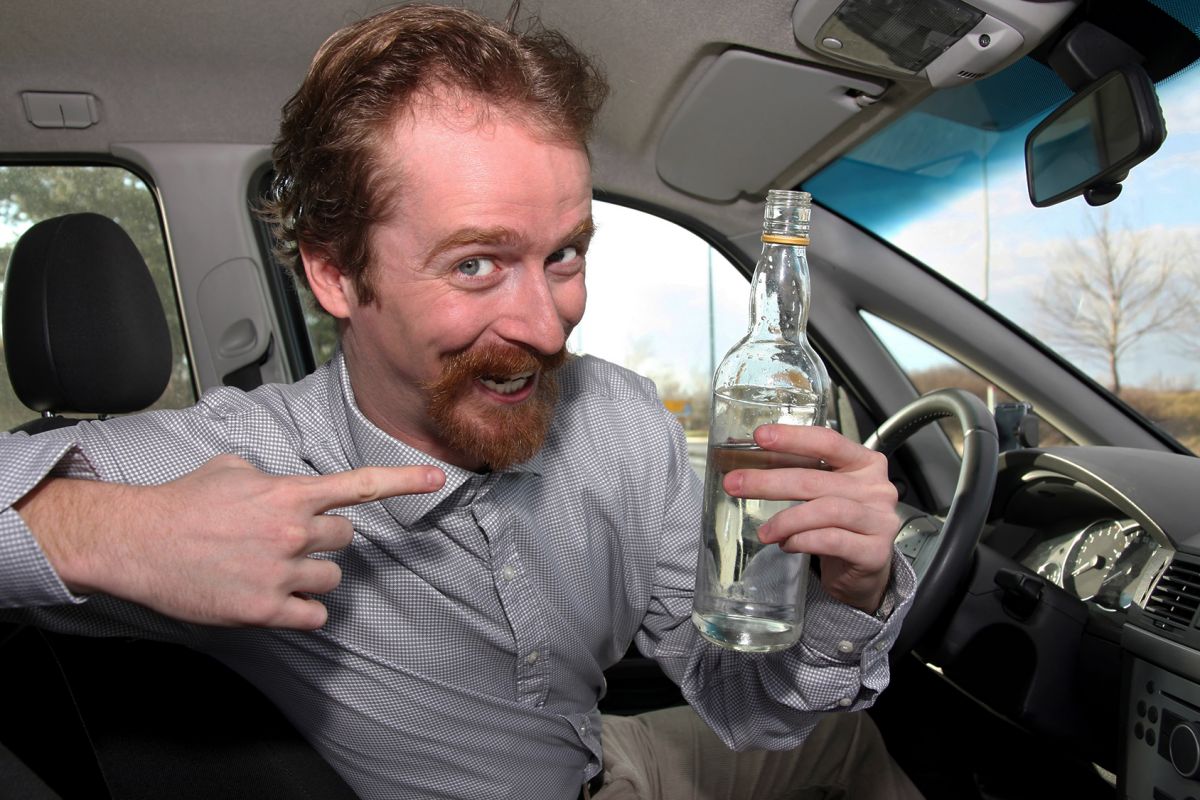 erheiterter mann im auto zeigt auf alkoholflasche