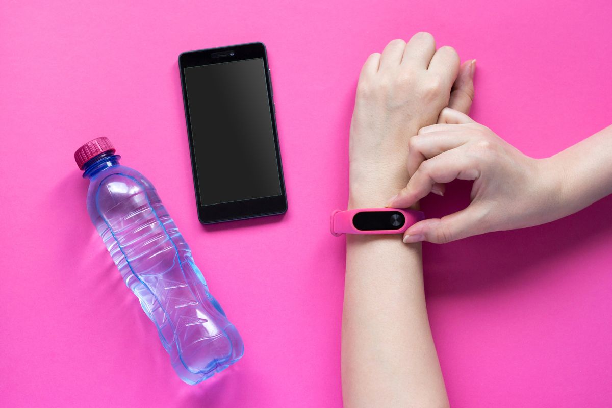 Schrittzähler an Handgelenk neben Smartphone und Wasserflasche auf pinkem Grund
