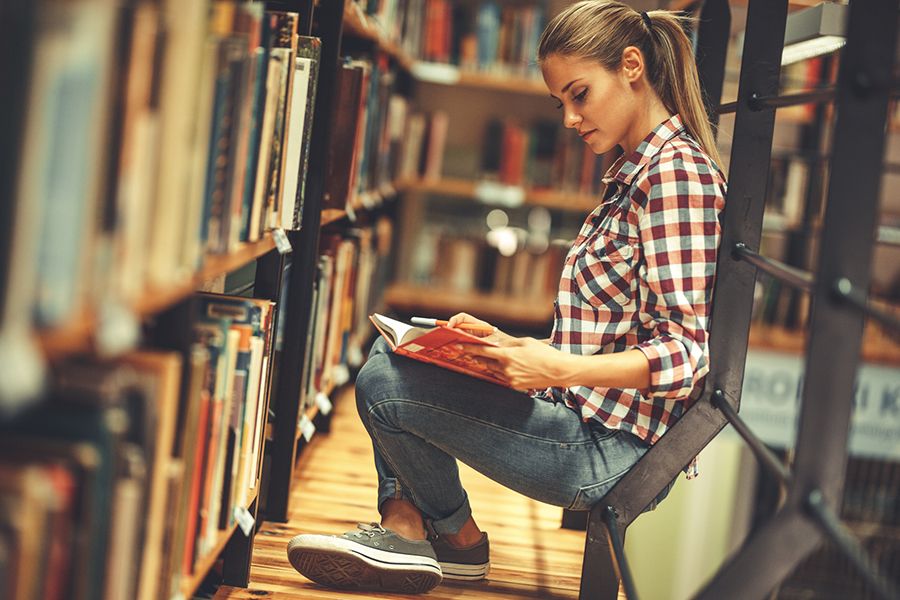 Online Sprachen lernen - Person sitzt in Bibliothek zwischen Buecheregalen