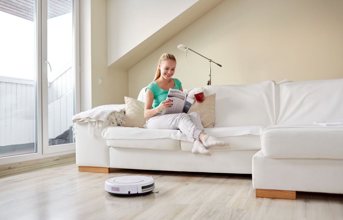 Frau liest auf Sofa, Staubsauger-Roboter saugt davor