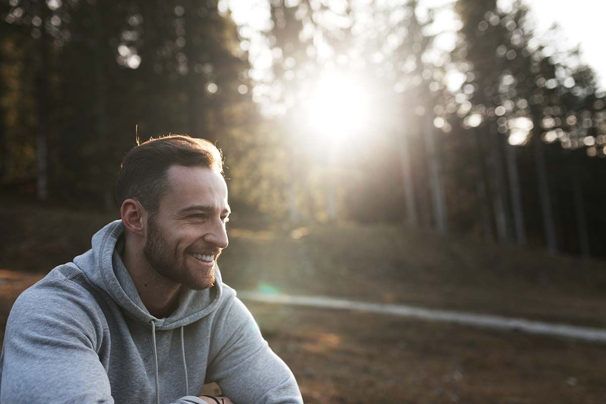 Junger Mann sitzt grinsend im Wald, während die Sonne untergeht
